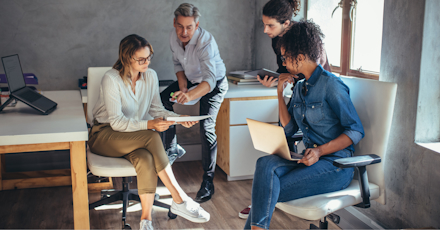 A group of four colleagues is engaged in a collaborative discussion in a modern office setting. One woman holds a document while another reviews it, with two other team members contributing to the conversation. Laptops and tablets are present, indicating a productive work session. The office environment is casual and bright, with natural light streaming through the windows, reflecting a focused and dynamic team effort.
