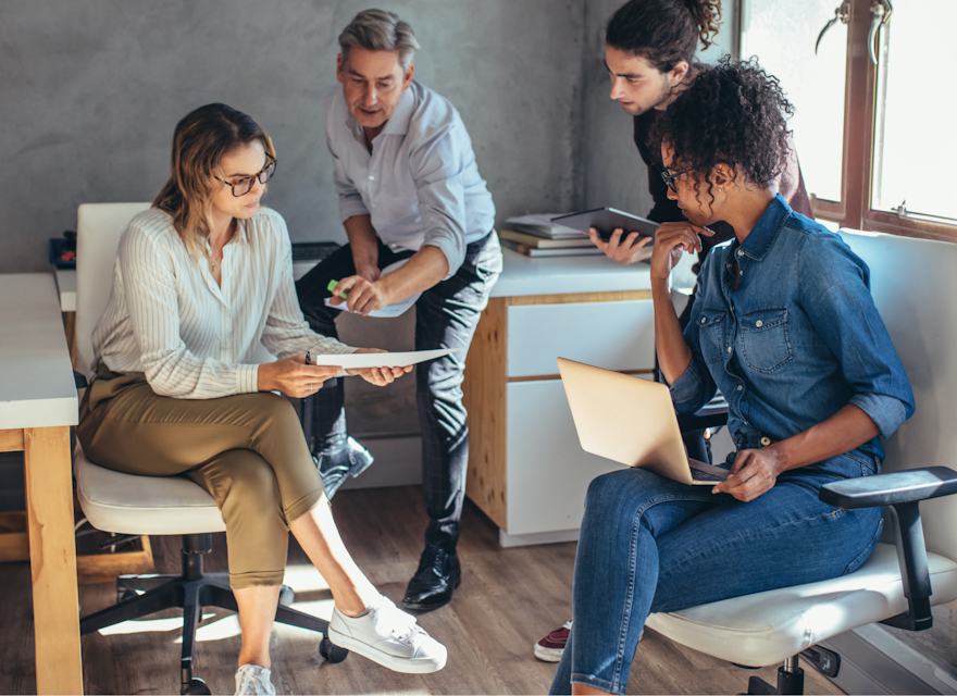A group of four colleagues is engaged in a collaborative discussion in a modern office setting. One woman holds a document while another reviews it, with two other team members contributing to the conversation. Laptops and tablets are present, indicating a productive work session. The office environment is casual and bright, with natural light streaming through the windows, reflecting a focused and dynamic team effort.