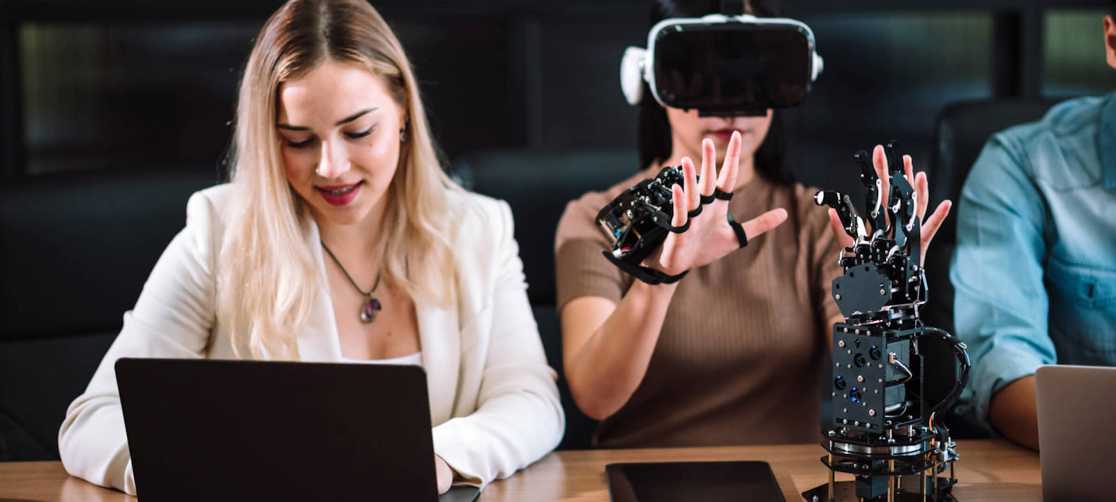 Woman using VR gloves and headset for robotics testing, while colleague works on laptop.