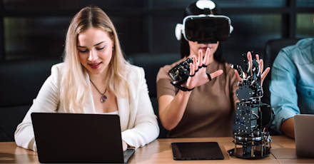 Woman using VR gloves and headset for robotics testing, while colleague works on laptop.