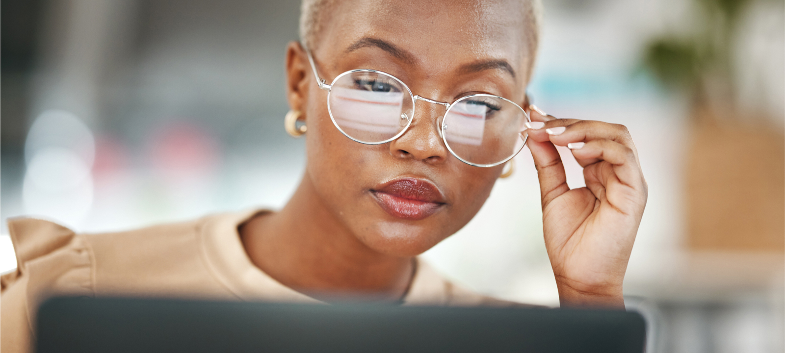 A close-up of a woman with short hair and large, round glasses, intently focused on her work. She is adjusting her glasses while looking at a computer screen, reflecting concentration and determination. The background is softly blurred, keeping the attention on her thoughtful expression and professional demeanor. The scene conveys a sense of focus and dedication in a modern, possibly office or remote work environment.