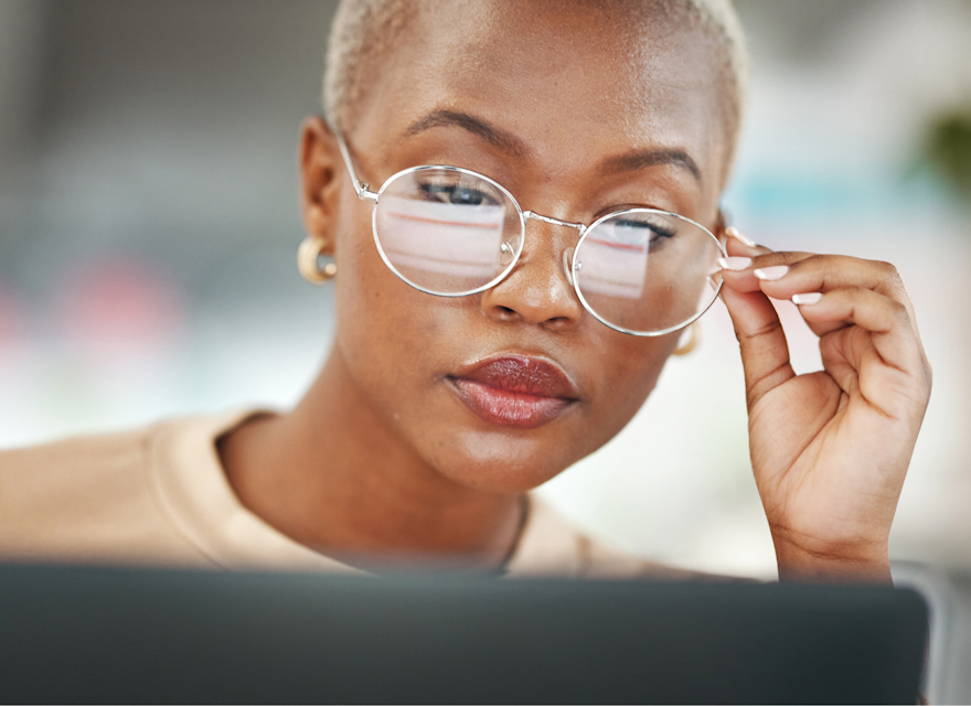 A close-up of a woman with short hair and large, round glasses, intently focused on her work. She is adjusting her glasses while looking at a computer screen, reflecting concentration and determination. The background is softly blurred, keeping the attention on her thoughtful expression and professional demeanor. The scene conveys a sense of focus and dedication in a modern, possibly office or remote work environment.