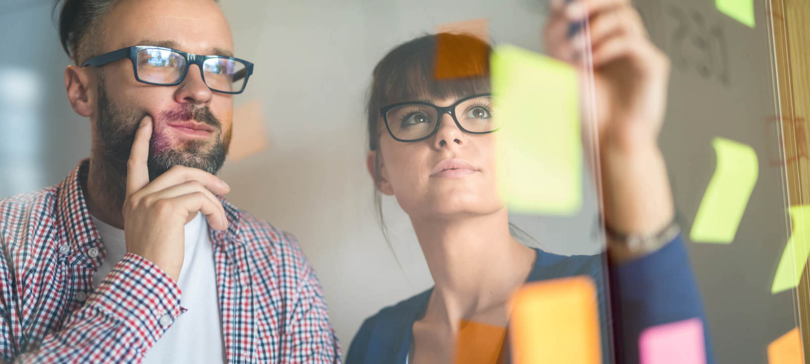 A man and woman, both wearing glasses, are brainstorming in front of a glass wall filled with colorful sticky notes. The man is deep in thought, while the woman is placing or adjusting a note. The scene captures the collaborative and creative process of problem-solving in a modern office environment. The vibrant notes and reflective glass suggest active planning and idea generation.