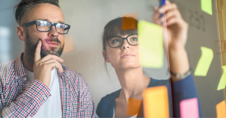 A man and woman, both wearing glasses, are brainstorming in front of a glass wall filled with colorful sticky notes. The man is deep in thought, while the woman is placing or adjusting a note. The scene captures the collaborative and creative process of problem-solving in a modern office environment. The vibrant notes and reflective glass suggest active planning and idea generation.