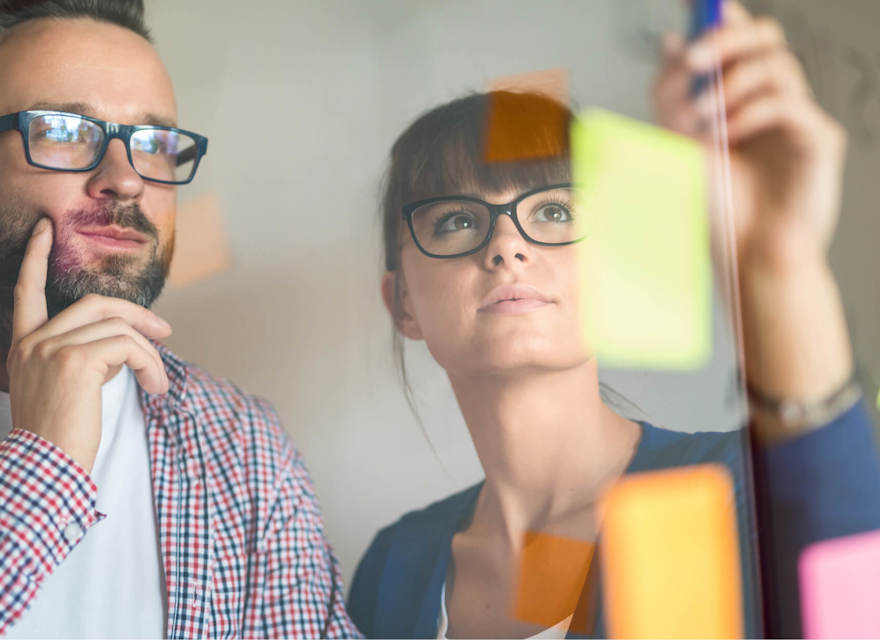 A man and woman, both wearing glasses, are brainstorming in front of a glass wall filled with colorful sticky notes. The man is deep in thought, while the woman is placing or adjusting a note. The scene captures the collaborative and creative process of problem-solving in a modern office environment. The vibrant notes and reflective glass suggest active planning and idea generation.