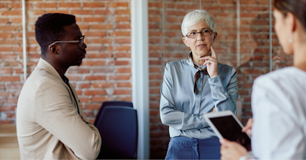 A diverse group of professionals engaged in a thoughtful discussion in a modern office setting. The group includes a senior woman with short white hair and glasses, who is speaking while holding her chin, and a younger man listening intently with his arms crossed. Another person, partially visible and holding a tablet, is involved in the conversation. The background features an exposed brick wall and glass partitions, giving the office a contemporary, open feel. The atmosphere suggests a strategic meeting or brainstorming session.