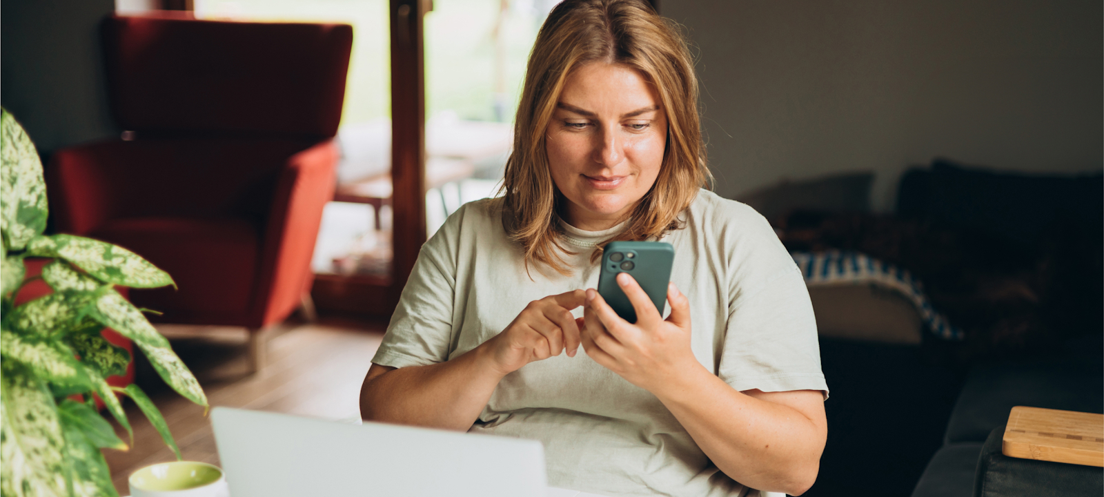 A woman is sitting at a table in a cozy, well-lit living space, using her smartphone. She appears content and focused, smiling slightly as she interacts with the device. A laptop is open on the table in front of her, alongside a coffee cup, suggesting she might be working or engaging in leisure activities. The background includes a red armchair, large windows allowing natural light, and a potted plant, contributing to the warm, inviting atmosphere of the room.