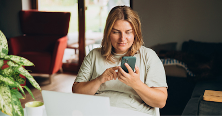 A woman is sitting at a table in a cozy, well-lit living space, using her smartphone. She appears content and focused, smiling slightly as she interacts with the device. A laptop is open on the table in front of her, alongside a coffee cup, suggesting she might be working or engaging in leisure activities. The background includes a red armchair, large windows allowing natural light, and a potted plant, contributing to the warm, inviting atmosphere of the room.