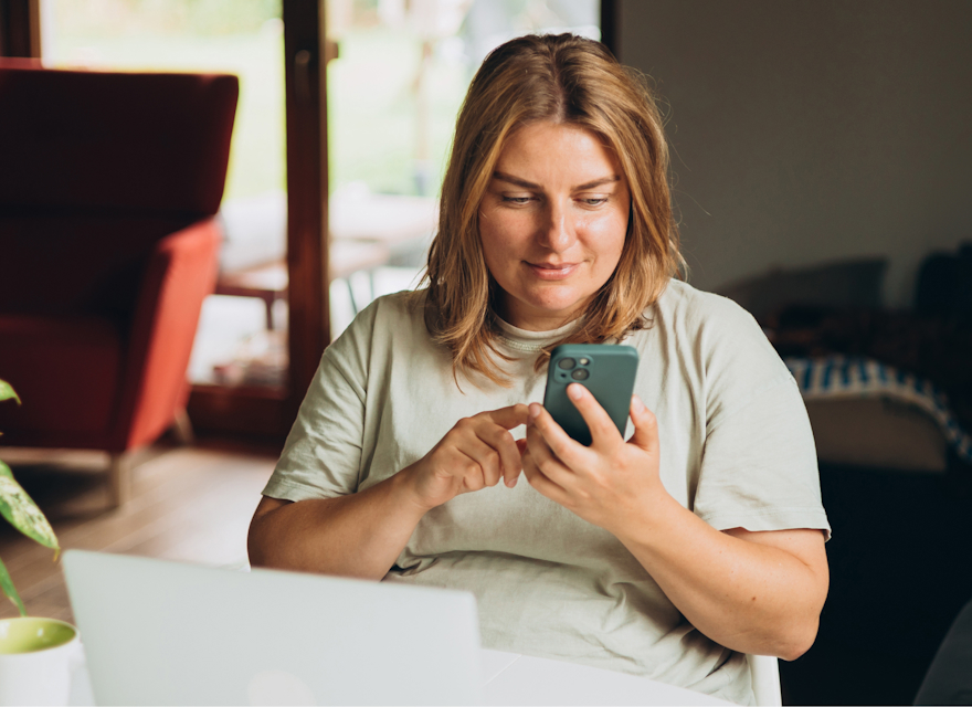 A woman is sitting at a table in a cozy, well-lit living space, using her smartphone. She appears content and focused, smiling slightly as she interacts with the device. A laptop is open on the table in front of her, alongside a coffee cup, suggesting she might be working or engaging in leisure activities. The background includes a red armchair, large windows allowing natural light, and a potted plant, contributing to the warm, inviting atmosphere of the room.