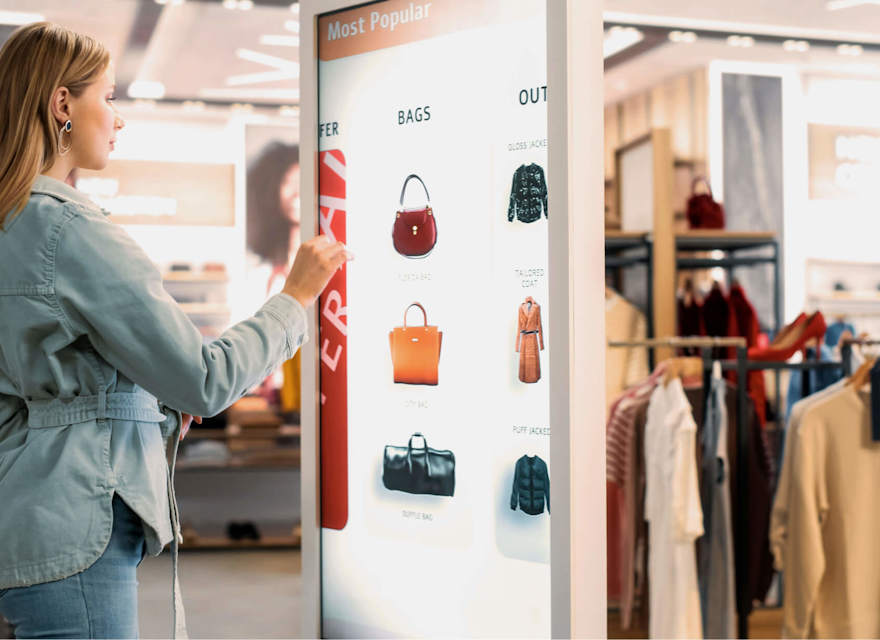 Woman using an interactive digital kiosk to browse handbags in a modern retail store.