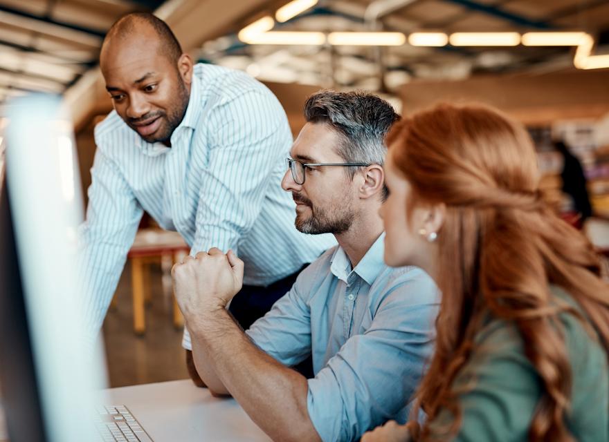 A diverse group of three colleagues collaborating at an office desk, with one person standing and two seated, focusing on a computer screen while discussing ideas.