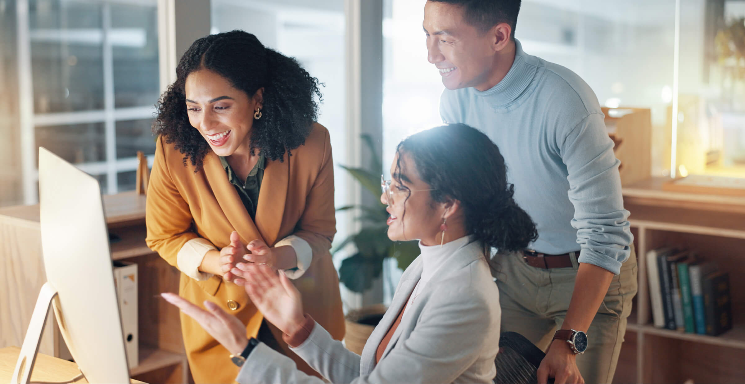 A group of three colleagues are gathered around a computer in a modern office, engaged in a collaborative discussion. One woman is seated, pointing at the screen while the other two, a man and a woman, stand beside her, smiling and interacting with the content on the monitor. The scene conveys a positive and energetic work environment, highlighting teamwork and collaboration. The office is bright and well-lit, with bookshelves and modern decor in the background.
