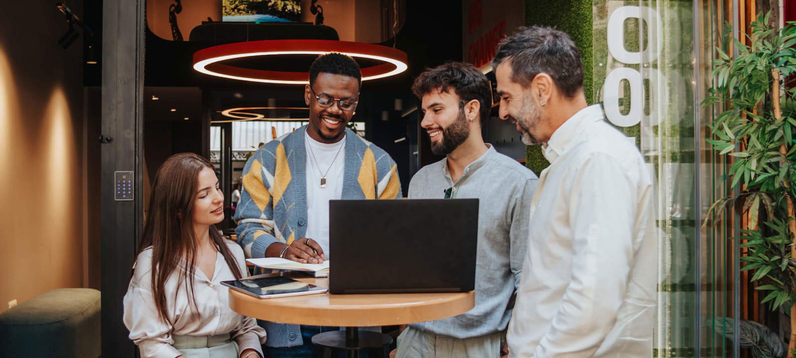 A diverse group of colleagues is gathered around a laptop during an outdoor meeting. The group consists of four men and one woman, all smiling and engaged in a collaborative discussion. They are standing and sitting around a high table in a modern outdoor space, with greenery and contemporary architecture in the background. The setting is casual and vibrant, reflecting a positive and creative work environment.
