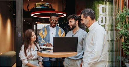 A diverse group of colleagues is gathered around a laptop during an outdoor meeting. The group consists of four men and one woman, all smiling and engaged in a collaborative discussion. They are standing and sitting around a high table in a modern outdoor space, with greenery and contemporary architecture in the background. The setting is casual and vibrant, reflecting a positive and creative work environment.