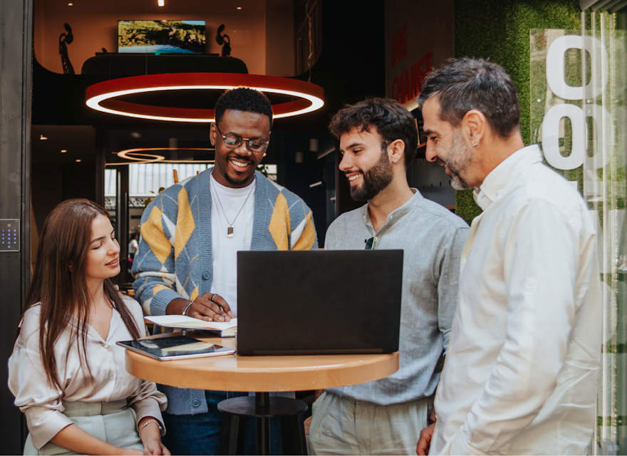 A diverse group of colleagues is gathered around a laptop during an outdoor meeting. The group consists of four men and one woman, all smiling and engaged in a collaborative discussion. They are standing and sitting around a high table in a modern outdoor space, with greenery and contemporary architecture in the background. The setting is casual and vibrant, reflecting a positive and creative work environment.