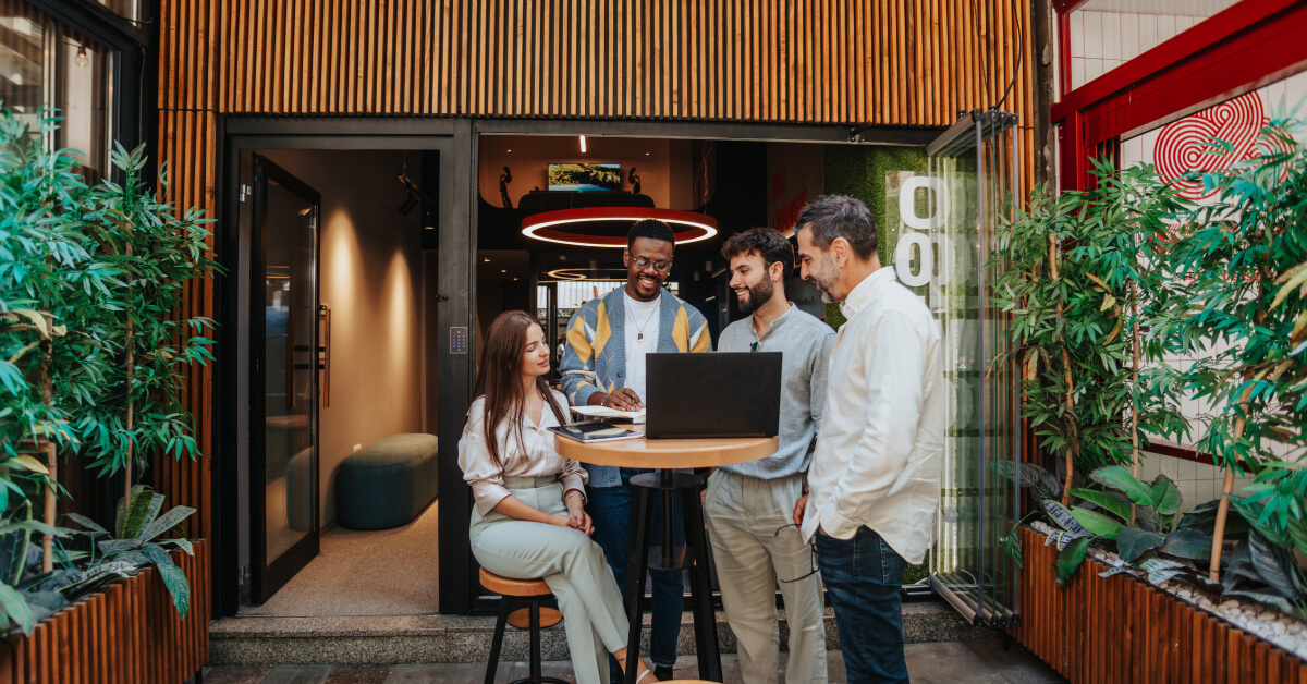 A diverse group of colleagues is gathered around a laptop during an outdoor meeting. The group consists of four men and one woman, all smiling and engaged in a collaborative discussion. They are standing and sitting around a high table in a modern outdoor space, with greenery and contemporary architecture in the background. The setting is casual and vibrant, reflecting a positive and creative work environment.