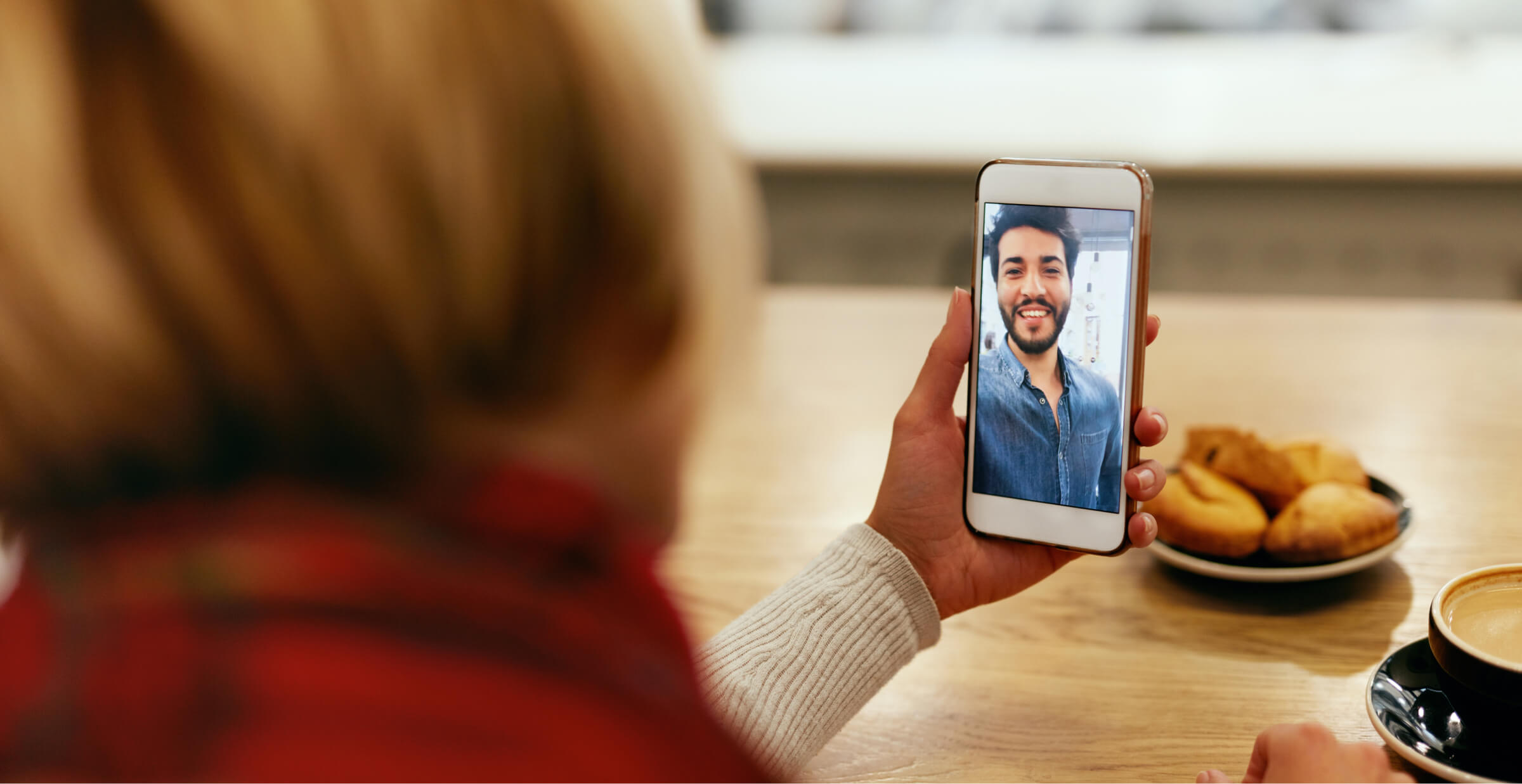 A person is sitting in a café, holding a smartphone while on a video call with a smiling man who appears on the screen. The table in front of the person has a cup of coffee and a plate of pastries, adding to the cozy atmosphere. The setting is warm and inviting, suggesting a relaxed and enjoyable conversation, possibly with a friend or loved one.