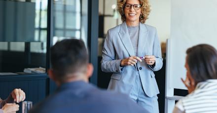 A businesswoman in a light gray suit smiles while leading a discussion with her team in a modern office setting.