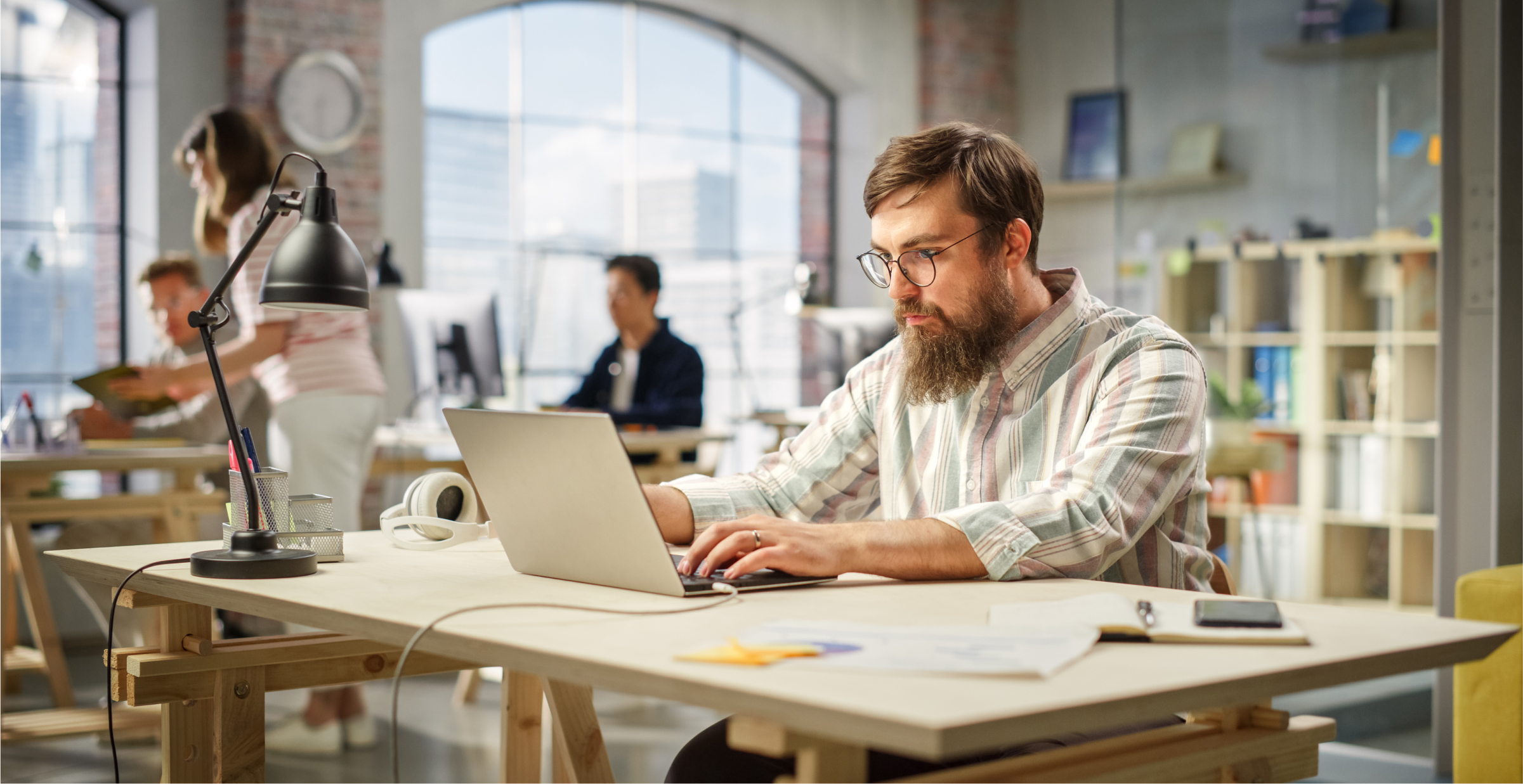 A man with a beard and glasses is working on a laptop at a wooden desk in a bright, modern office. The workspace features large windows with city views, allowing natural light to fill the room. The man appears focused, surrounded by office supplies, including a desk lamp, headphones, and a smartphone. In the background, other colleagues are engaged in work, contributing to a busy yet collaborative atmosphere. The office setting is open and contemporary, with a mix of industrial and modern design elements.