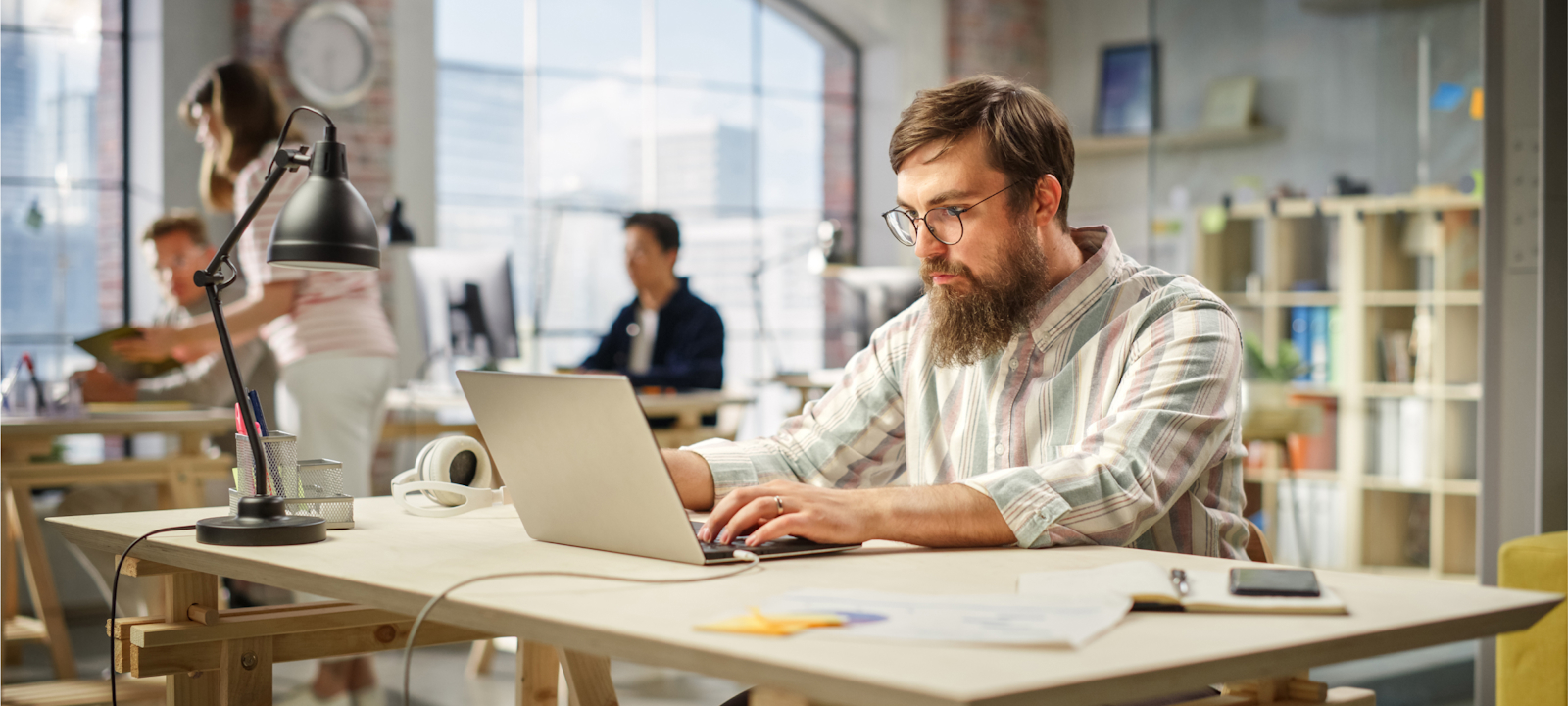 A man with a beard and glasses is working on a laptop at a wooden desk in a bright, modern office. The workspace features large windows with city views, allowing natural light to fill the room. The man appears focused, surrounded by office supplies, including a desk lamp, headphones, and a smartphone. In the background, other colleagues are engaged in work, contributing to a busy yet collaborative atmosphere. The office setting is open and contemporary, with a mix of industrial and modern design elements.