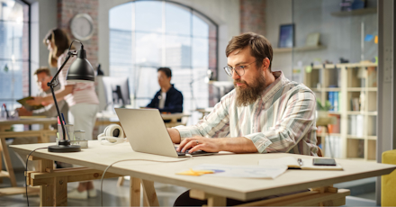 A man with a beard and glasses is working on a laptop at a wooden desk in a bright, modern office. The workspace features large windows with city views, allowing natural light to fill the room. The man appears focused, surrounded by office supplies, including a desk lamp, headphones, and a smartphone. In the background, other colleagues are engaged in work, contributing to a busy yet collaborative atmosphere. The office setting is open and contemporary, with a mix of industrial and modern design elements.