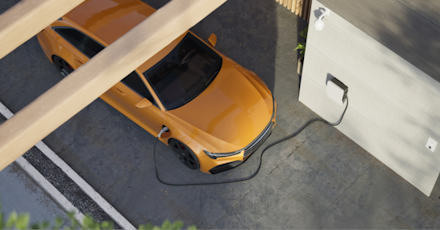 A top-down view of an orange electric car charging at a home charging station. The car is parked in a driveway, connected to a wall-mounted charging unit. The setting is modern and clean, with part of the car shaded by a wooden structure. The image highlights sustainability and the use of electric vehicles in residential environments.
