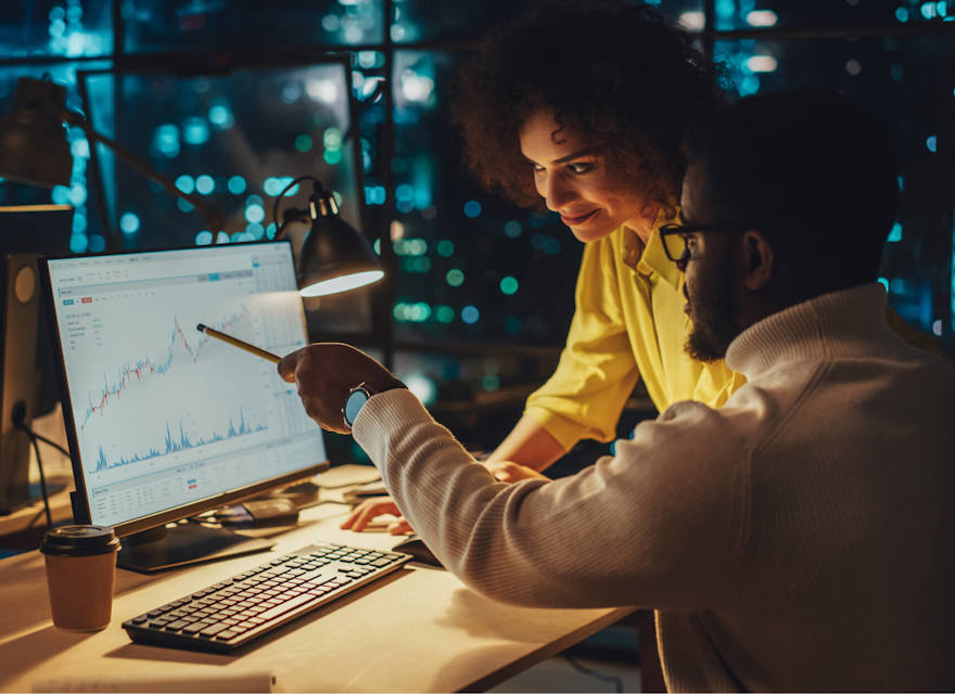 Team collaborating on financial analysis in a dimly lit office with computer screens displaying data charts.