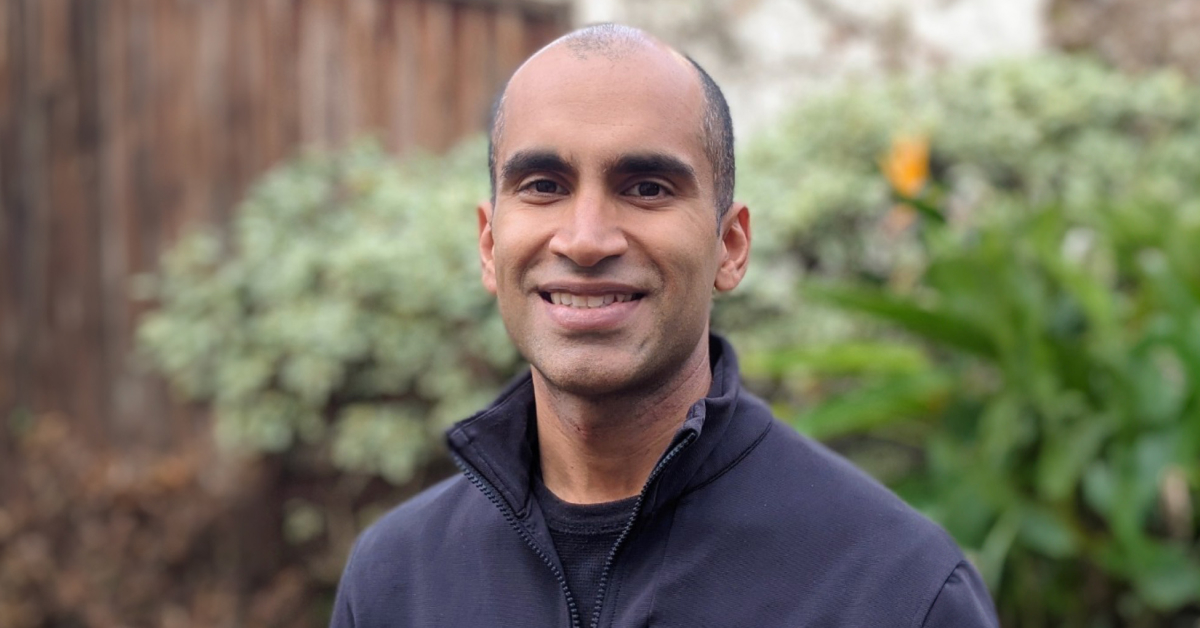 This image shows Nitin Garg, standing outdoors in front of a blurred background featuring green plants and a wooden fence. He has a closely shaved head, dark eyebrows, and a medium-dark complexion. He is wearing a dark-colored jacket or zip-up sweater. The lighting is soft and natural, suggesting the photo was taken during the daytime.
