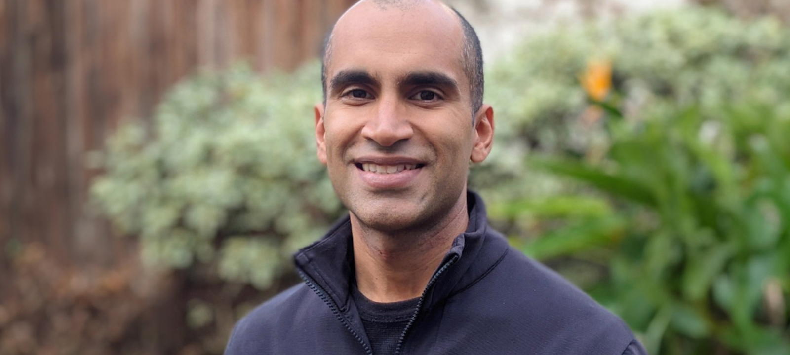 This image shows Nitin Garg, standing outdoors in front of a blurred background featuring green plants and a wooden fence. He has a closely shaved head, dark eyebrows, and a medium-dark complexion. He is wearing a dark-colored jacket or zip-up sweater. The lighting is soft and natural, suggesting the photo was taken during the daytime.