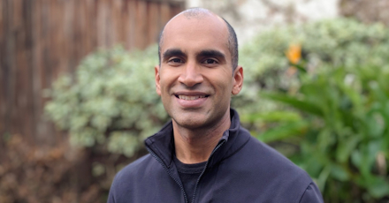 This image shows Nitin Garg, standing outdoors in front of a blurred background featuring green plants and a wooden fence. He has a closely shaved head, dark eyebrows, and a medium-dark complexion. He is wearing a dark-colored jacket or zip-up sweater. The lighting is soft and natural, suggesting the photo was taken during the daytime.