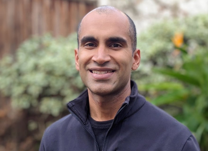 This image shows Nitin Garg, standing outdoors in front of a blurred background featuring green plants and a wooden fence. He has a closely shaved head, dark eyebrows, and a medium-dark complexion. He is wearing a dark-colored jacket or zip-up sweater. The lighting is soft and natural, suggesting the photo was taken during the daytime.