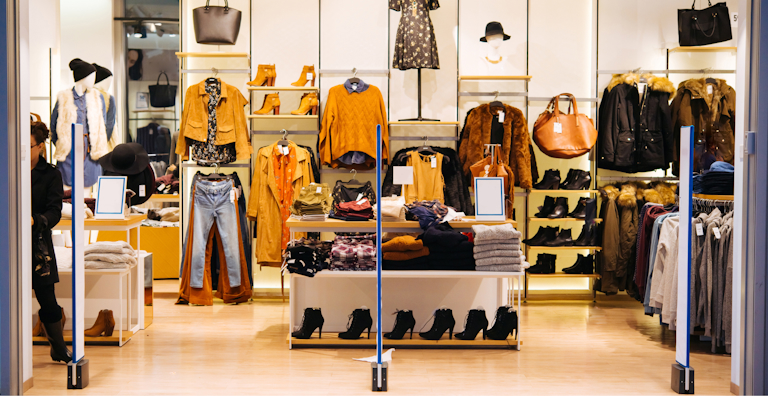 A well-organized clothing store display featuring a variety of stylish outfits and accessories. The display includes mannequins dressed in autumn and winter attire, such as a faux fur vest, a suede jacket, and a floral dress. Shelves and racks are stocked with folded sweaters, denim jeans, leather boots, handbags, and hats. The store has a warm, inviting atmosphere with soft lighting that highlights the earthy tones of the clothing. The overall setup is visually appealing, encouraging customers to browse and explore the fashion options.