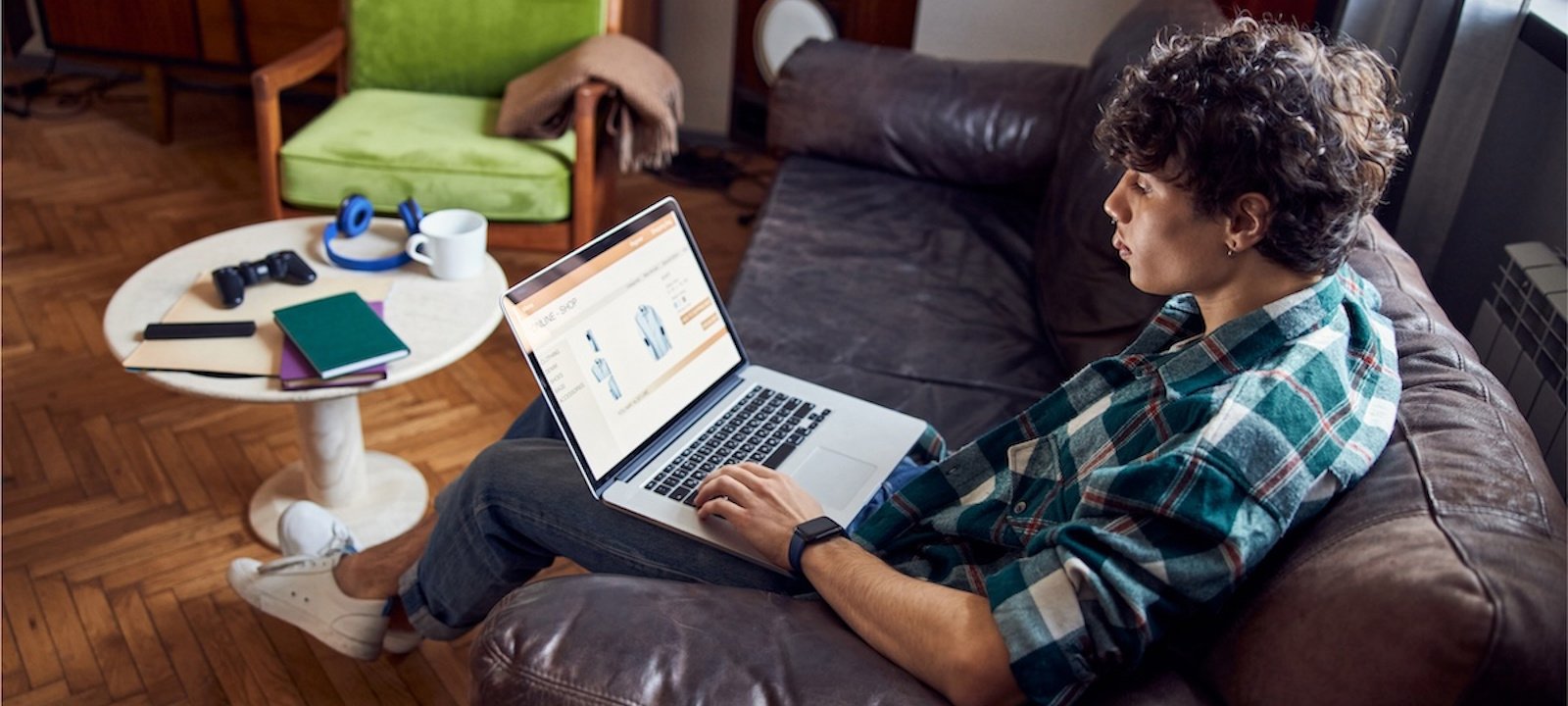 A young man with curly hair is sitting on a brown leather couch in a cozy living room, browsing an online store on his laptop. He is wearing a green and white plaid shirt, blue jeans, and white sneakers. A round white coffee table in front of him holds a gaming controller, notebooks, and a pair of headphones. The room has wooden flooring, a green armchair with a blanket draped over it, and warm lighting, creating a comfortable atmosphere.