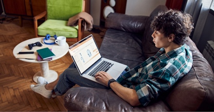 A young man with curly hair is sitting on a brown leather couch in a cozy living room, browsing an online store on his laptop. He is wearing a green and white plaid shirt, blue jeans, and white sneakers. A round white coffee table in front of him holds a gaming controller, notebooks, and a pair of headphones. The room has wooden flooring, a green armchair with a blanket draped over it, and warm lighting, creating a comfortable atmosphere.
