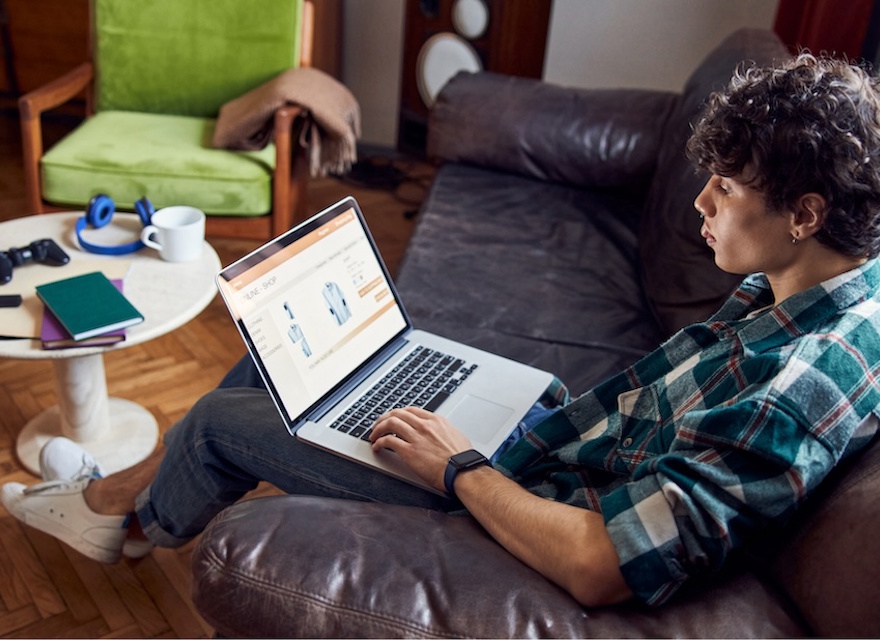 A young man with curly hair is sitting on a brown leather couch in a cozy living room, browsing an online store on his laptop. He is wearing a green and white plaid shirt, blue jeans, and white sneakers. A round white coffee table in front of him holds a gaming controller, notebooks, and a pair of headphones. The room has wooden flooring, a green armchair with a blanket draped over it, and warm lighting, creating a comfortable atmosphere.