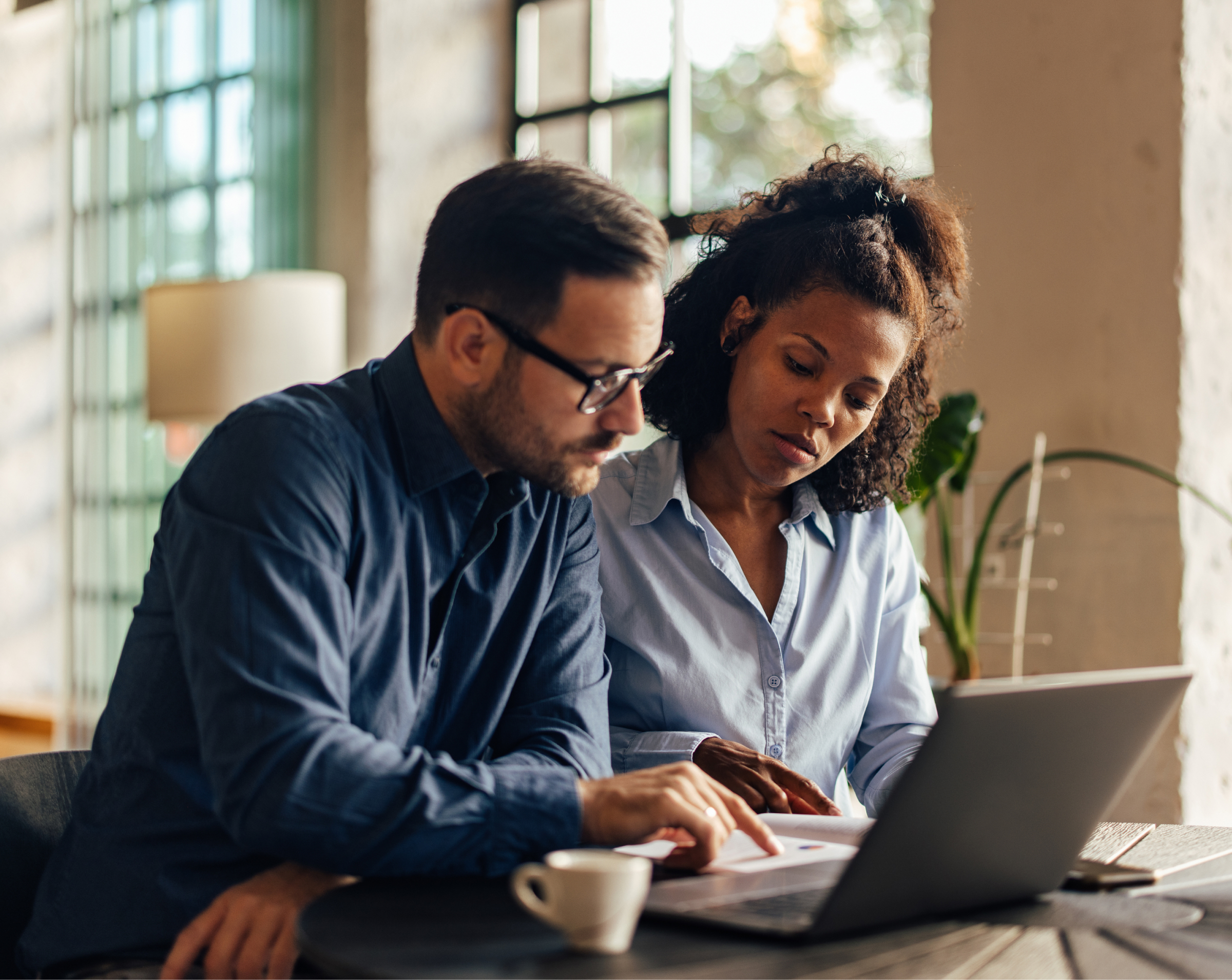 Two professionals collaborating at a table, reviewing documents on a laptop in a well-lit modern office space.