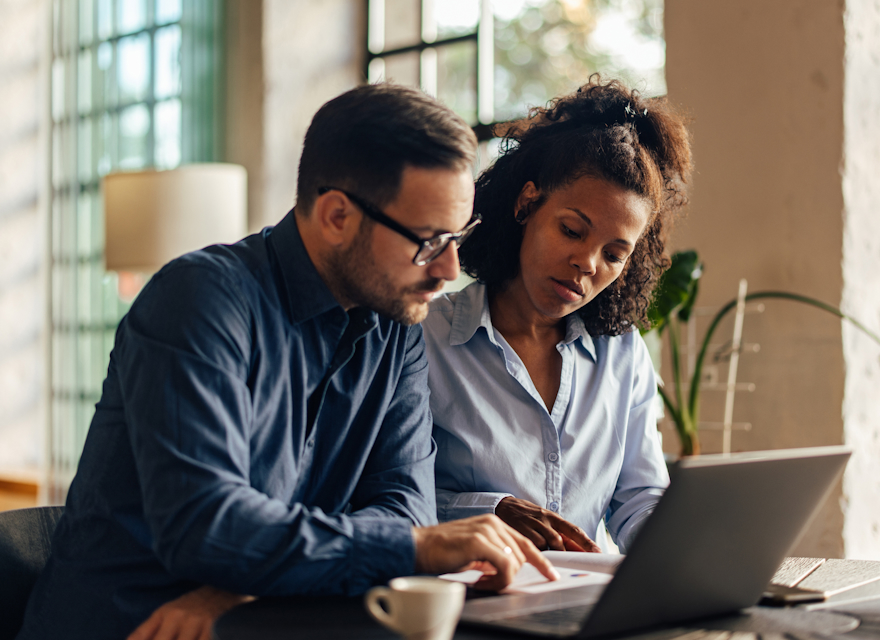 Two professionals collaborating at a table, reviewing documents on a laptop in a well-lit modern office space.