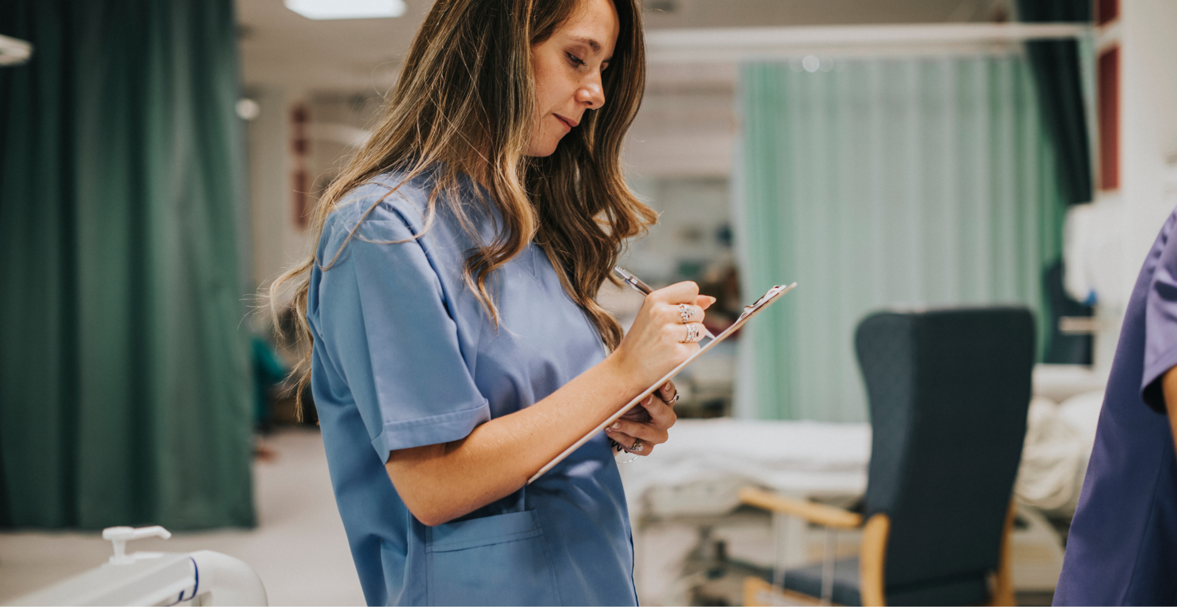 A nurse in a blue uniform writing notes on a clipboard in a hospital ward.
