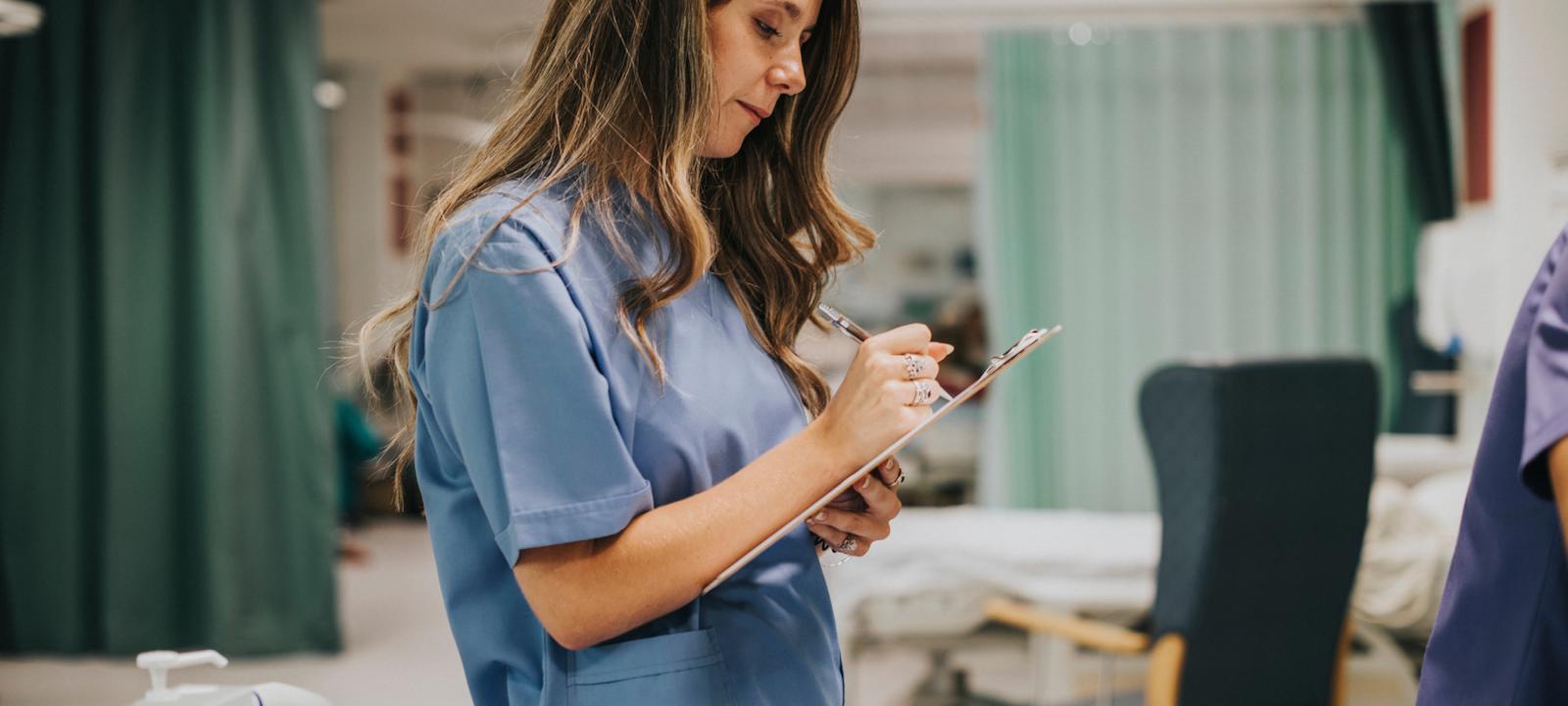 A nurse in a blue uniform writing notes on a clipboard in a hospital ward.