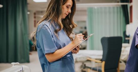 A nurse in a blue uniform writing notes on a clipboard in a hospital ward.