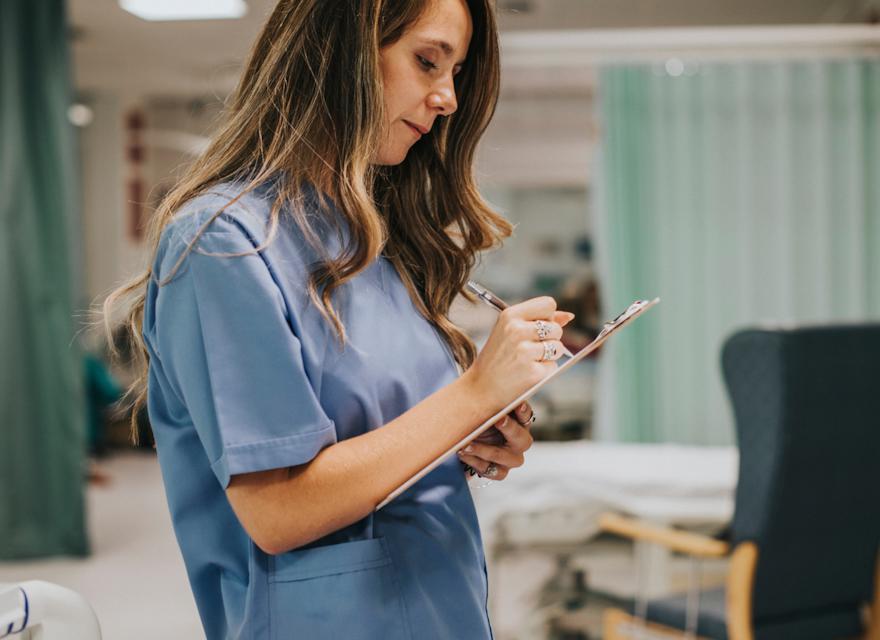 A nurse in a blue uniform writing notes on a clipboard in a hospital ward.