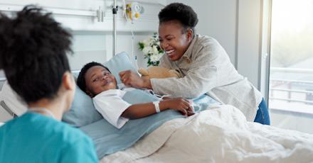 A cheerful hospital scene with a young patient in bed, smiling alongside a parent and a healthcare professional nearby.
