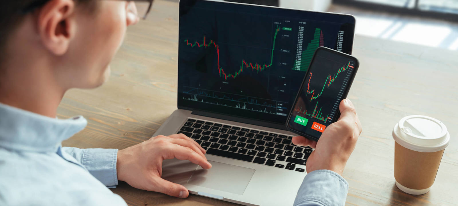 A man is sitting at a desk using both a laptop and a smartphone to monitor stock trading charts. The laptop and phone screens display live financial data with price charts and buy/sell options. A takeaway coffee cup is placed on the desk next to him, indicating a focused and dynamic work environment. The image conveys modern investment management and the use of technology in financial trading.