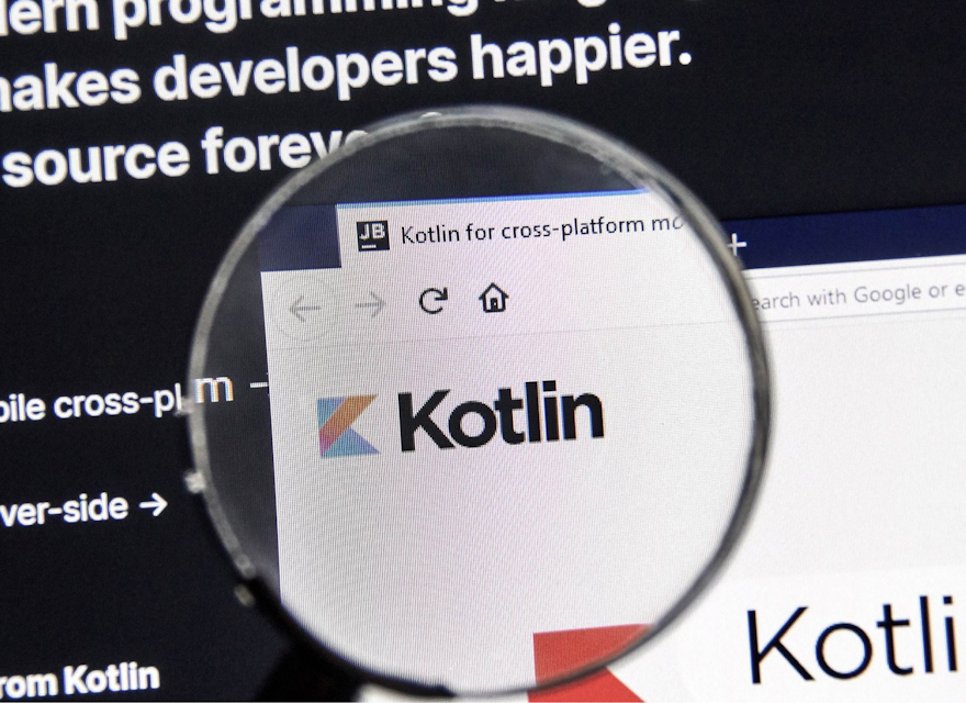 A close-up of a computer screen showing the Kotlin programming language website through a magnifying glass. The screen highlights that Kotlin is a modern, open-source programming language, with emphasis on mobile cross-platform and server-side development. The magnified section focuses on the Kotlin logo, reinforcing its relevance in the developer community. The image represents the importance of Kotlin in modern software development.