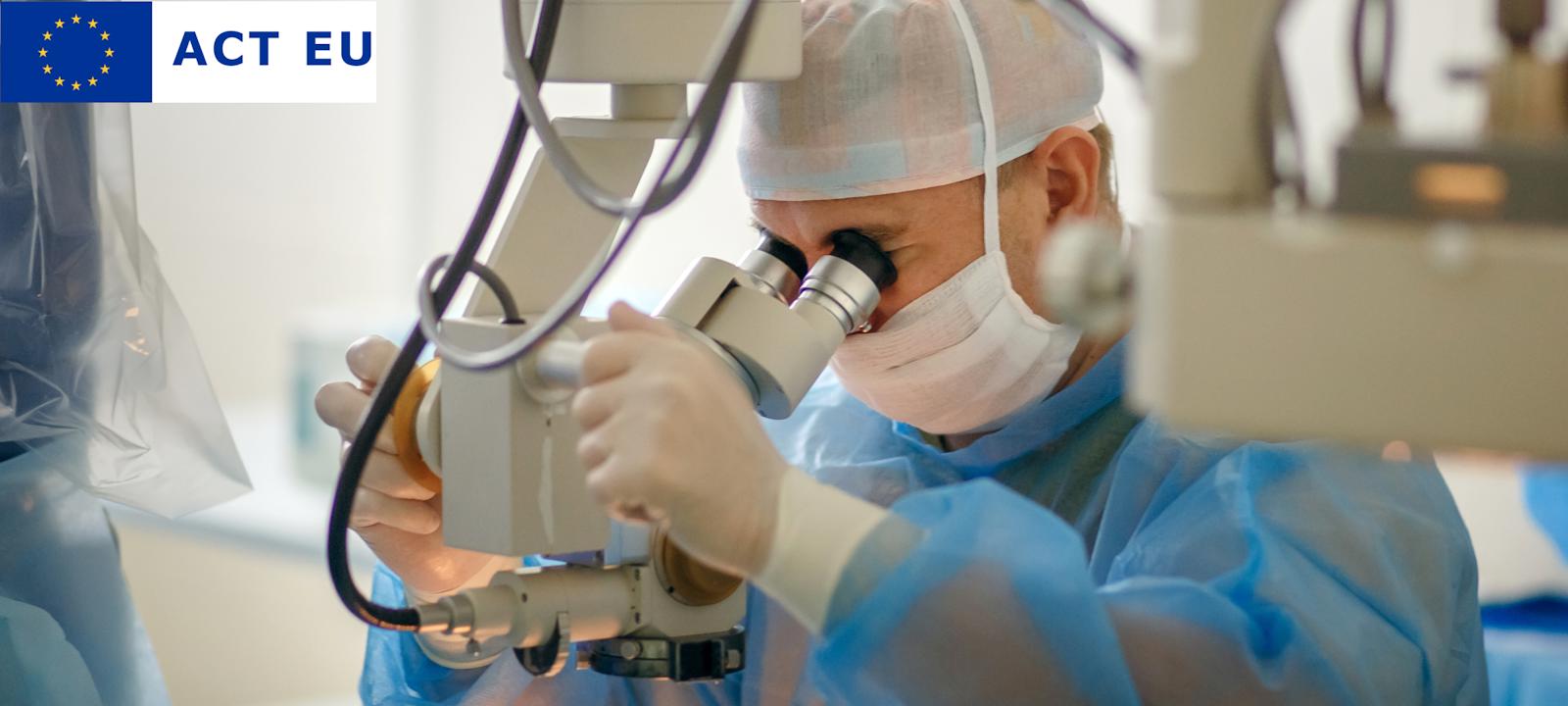 A surgeon wearing a blue surgical gown, gloves, a face mask, and a hair cap is using a high-precision operating microscope during a medical procedure. The sterile environment and advanced equipment indicate a surgical setting, likely for ophthalmic or microsurgery.