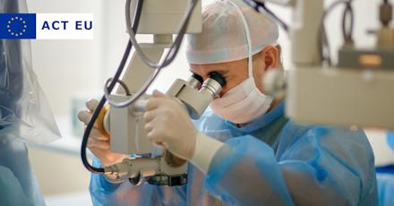 A surgeon wearing a blue surgical gown, gloves, a face mask, and a hair cap is using a high-precision operating microscope during a medical procedure. The sterile environment and advanced equipment indicate a surgical setting, likely for ophthalmic or microsurgery.