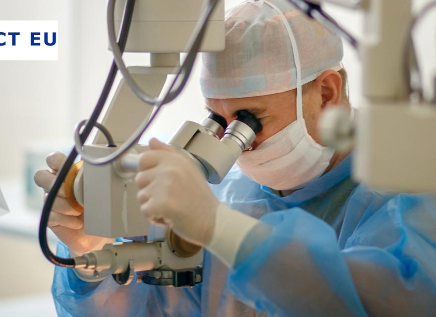 A surgeon wearing a blue surgical gown, gloves, a face mask, and a hair cap is using a high-precision operating microscope during a medical procedure. The sterile environment and advanced equipment indicate a surgical setting, likely for ophthalmic or microsurgery.