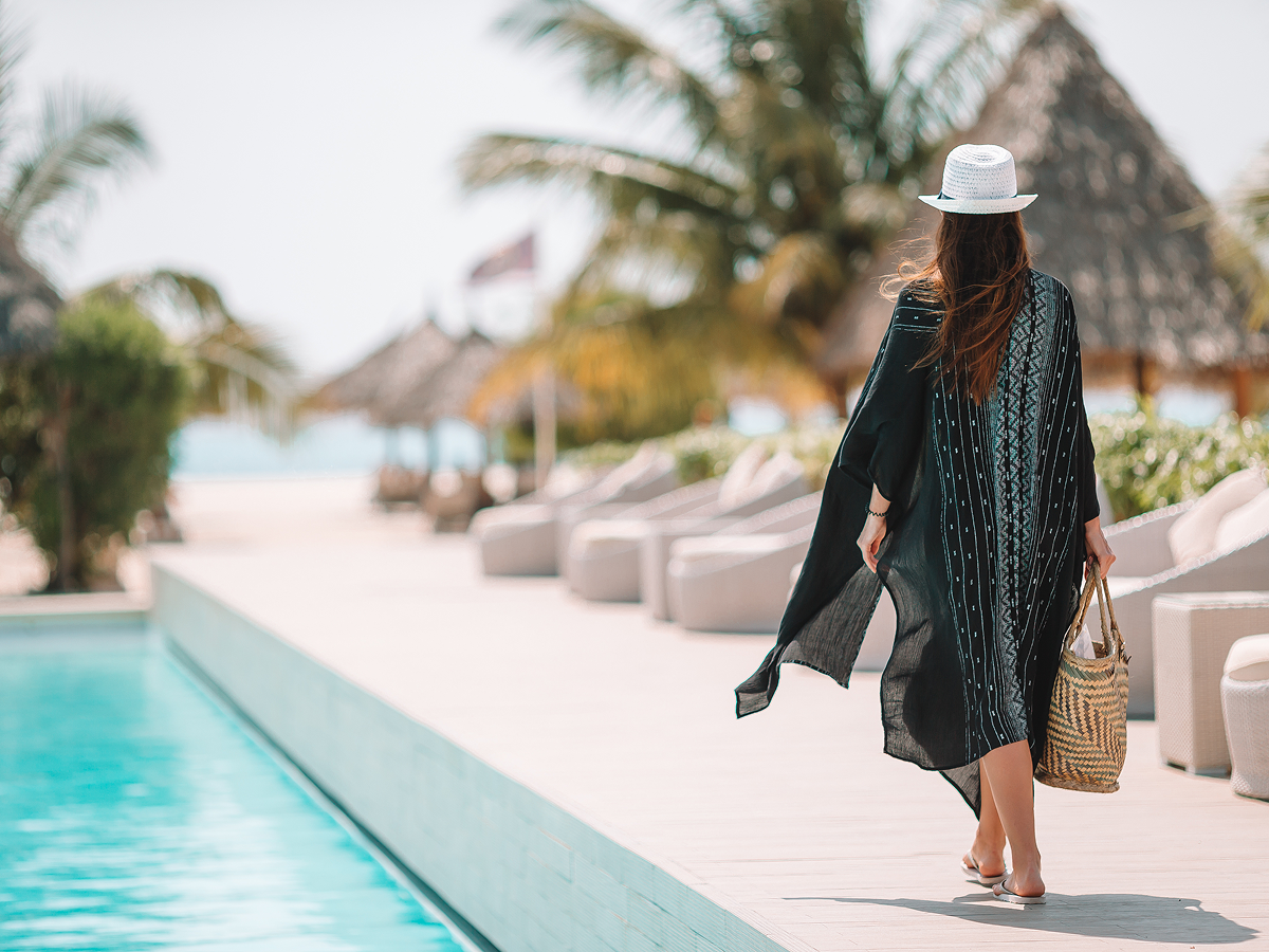 A woman in a stylish hat and dress strolls by the pool, enjoying a sunny day