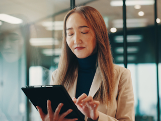 A woman in a business suit holds a tablet, appearing focused and engaged in a professional setting.