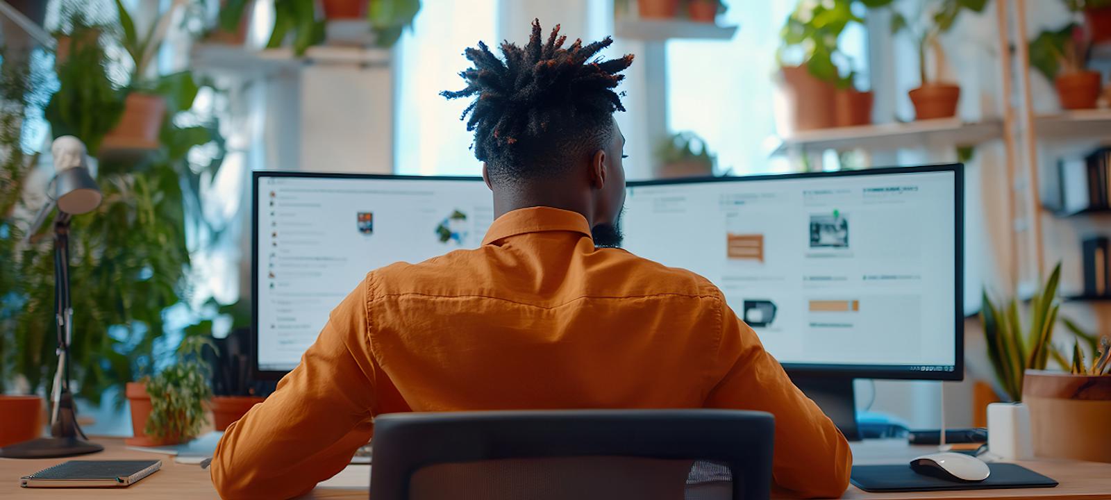 A man sits at a desk, focused on two computer screens displaying various work tasks.